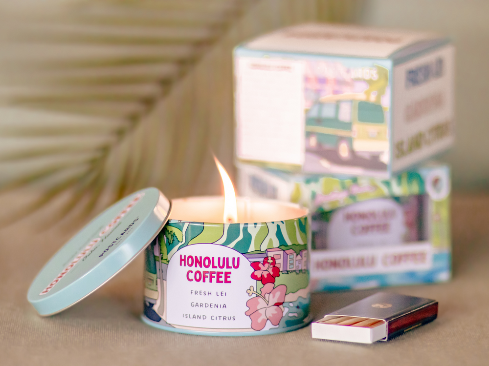Lit Postcards Candle with fresh lei, gardenia, and island citrus fragrances. Next to a box of matches and packaging. 