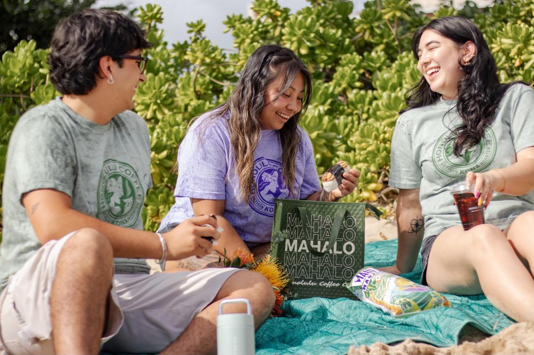 Three people sitting on a blanket outdoors with a 'Mahalo' sign and coffee bags.