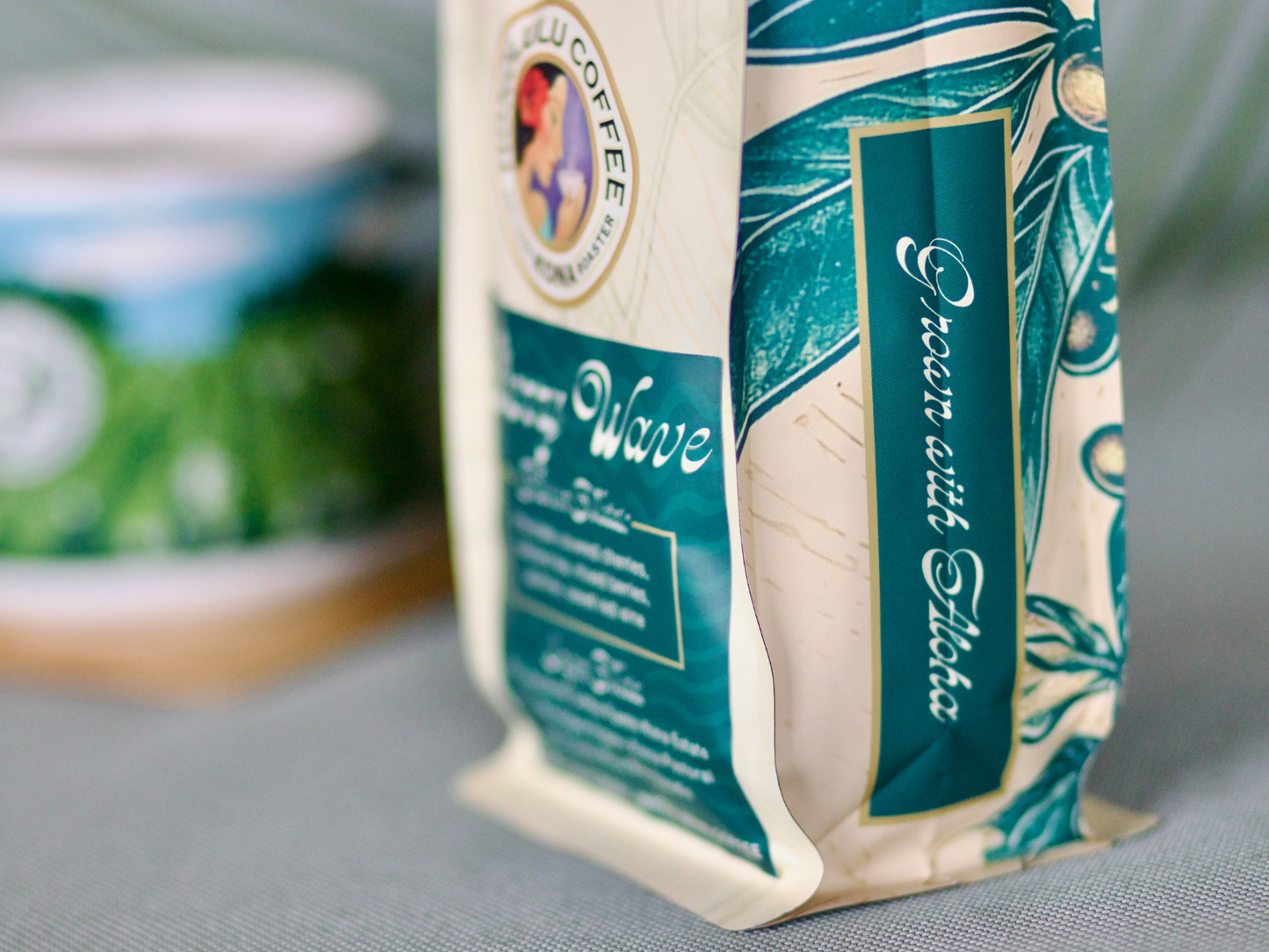 
                  
                    Details of the new Cherry Wave coffee blend bag. Teal coffee cherries with hints of gold. "Grown with Aloha" written on the sides. 
                  
                