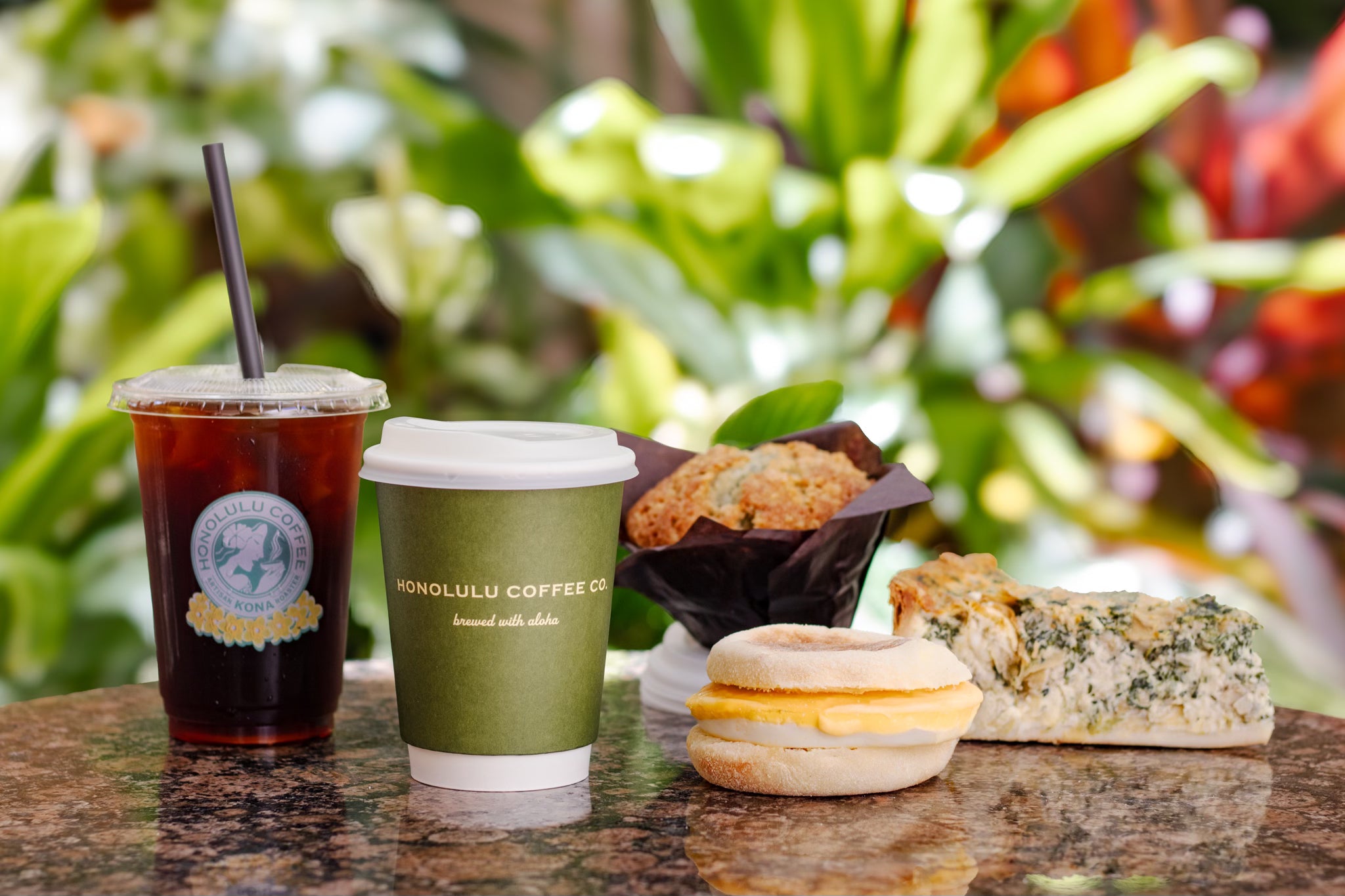 Two coffee drinks and a small assortment of pastries on a table with a blurred green outdoor background.