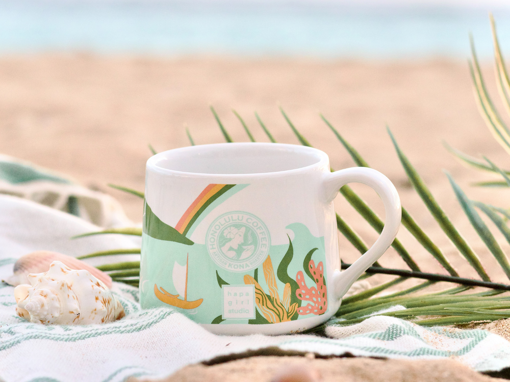 A mug on a beach towel with seashells on it.