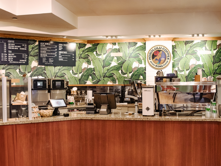 Coffee shop interior with a counter, green leafy wall design, and Honolulu Coffee logo.