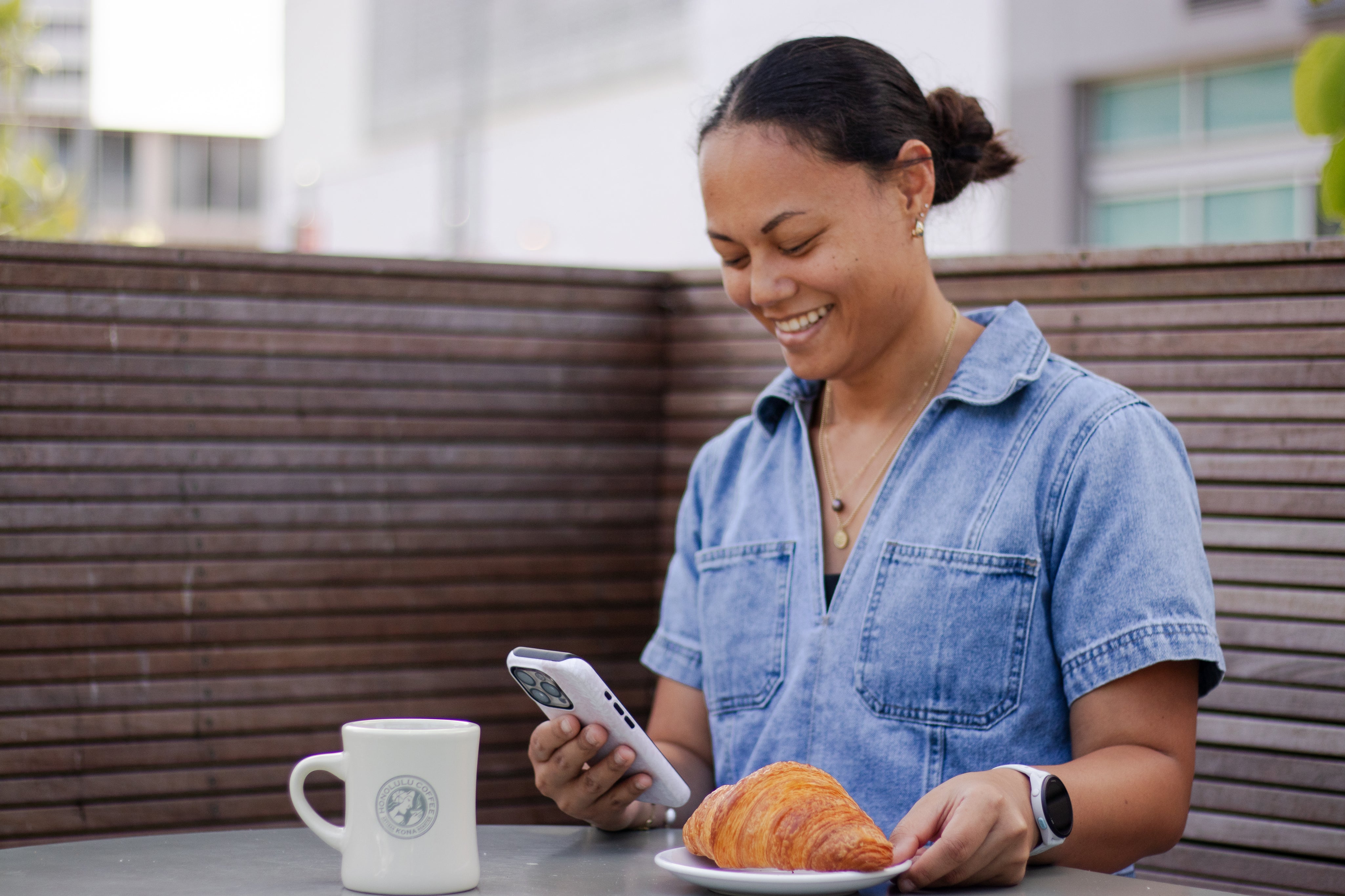 Woman using a smartphone with a croissant and coffee at an outdoor table.