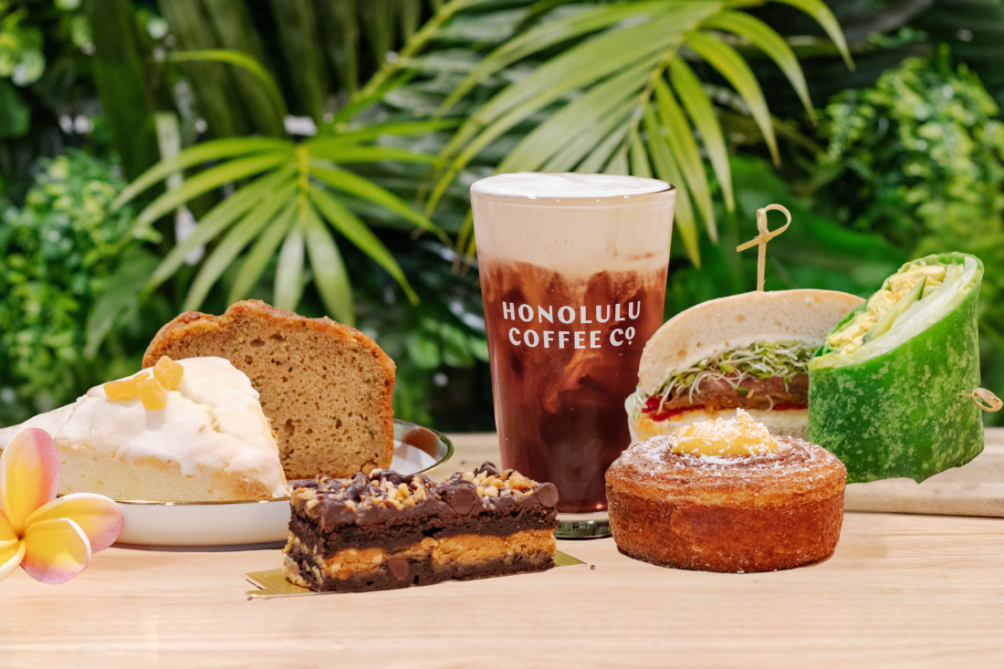 Assorted pastries and a Honolulu Coffee Co. iced coffee on a wooden table with greenery in the background.